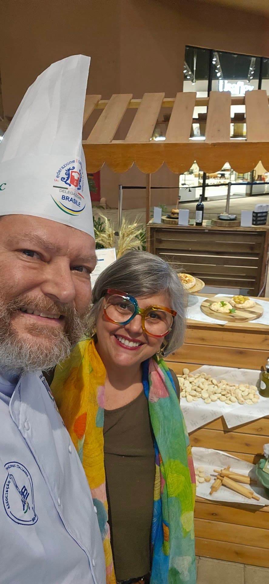 Chef Marcelo Lampe usando a toque branca da Federazione Italiana Cuochi, Delegazione Brasile, e dólmã com o brasão da associação, ao lado de Daniella, em um evento gastronômico com bancadas de massas e pães ao fundo.