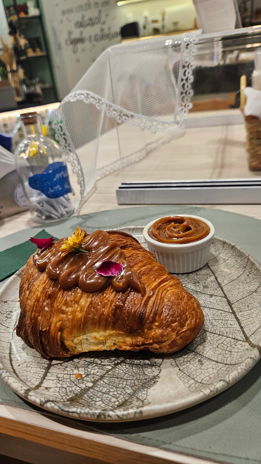 Croissant folhado coberto com doce de leite e flores comestíveis amarelas e roxas, servido em prato artesanal com um ramequim de doce de leite ao lado. Ao fundo, o vidro de bilhetes da casa.