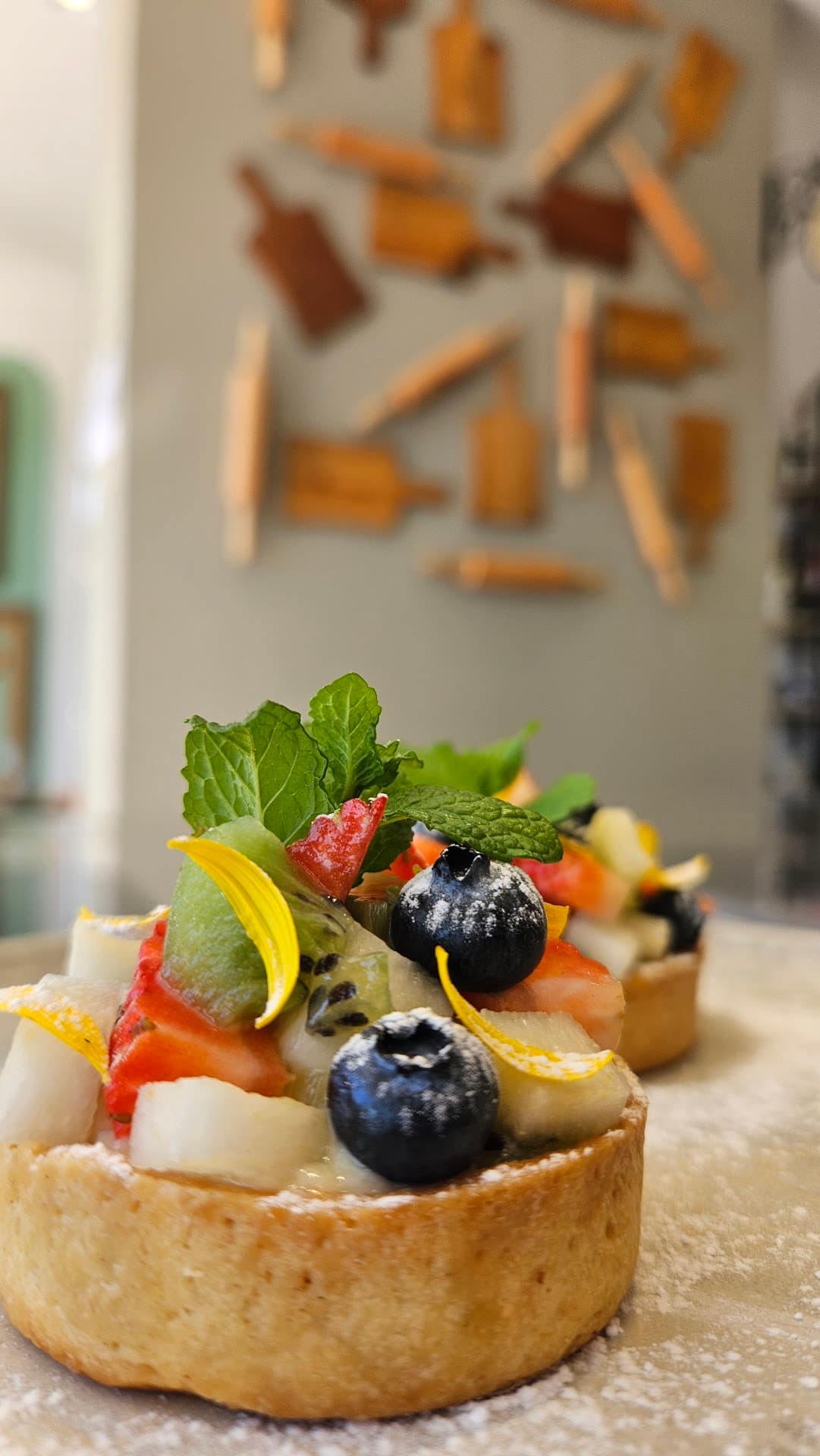 Tartelete de frutas com morango, kiwi, mirtilo, pétalas amarelas e hortelã, fotografada de perto sobre a bancada polvilhada de açúcar.