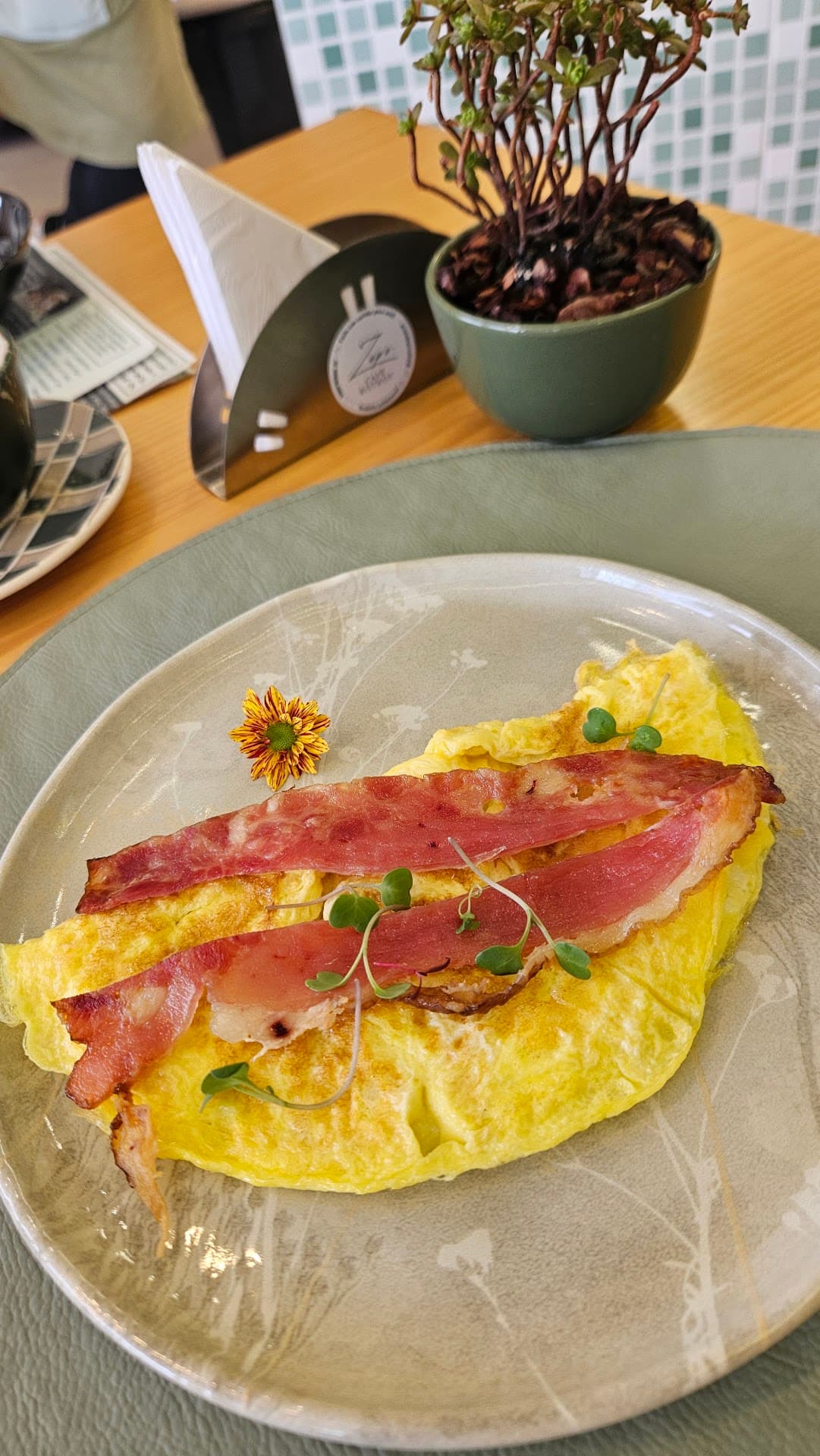 Omelete com bacon e brotos em prato artesanal sobre mesa de madeira clara, com porta-guardanapos gravado com o logo do Zepe e um vaso verde com planta ao fundo, diante da parede de pastilhas verdes.