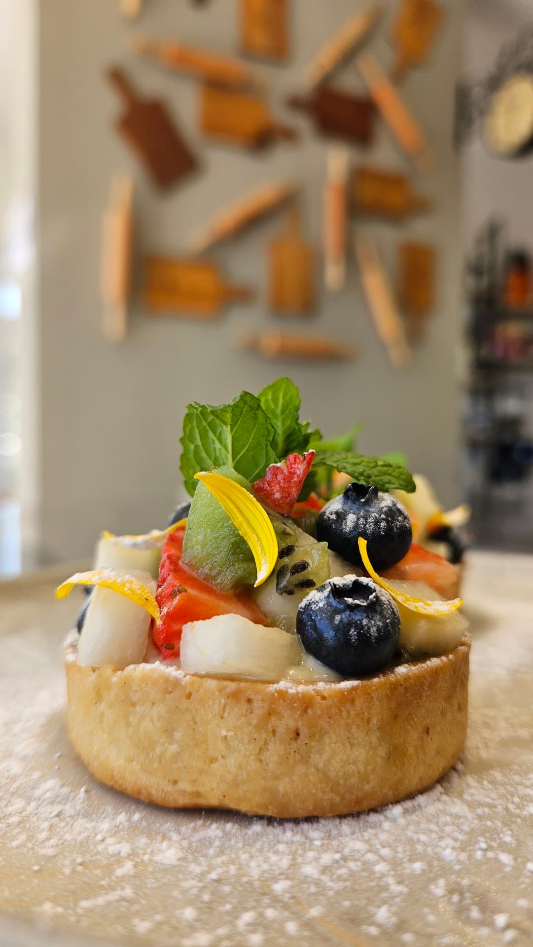 Tartelete de frutas frescas (morango, kiwi, mirtilo e hortelã) sobre creme, polvilhada com açúcar, tendo ao fundo, desfocada, a parede de rolos de macarrão e tábuas de madeira do salão do Zepe.